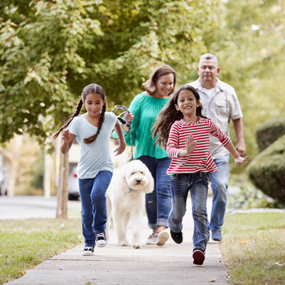 Smiling family of four walking down sidewalk with two young daughters running in front of parents walking dog Smiling family of four walking down sidewalk with two young daughters running in front of parents walking dog