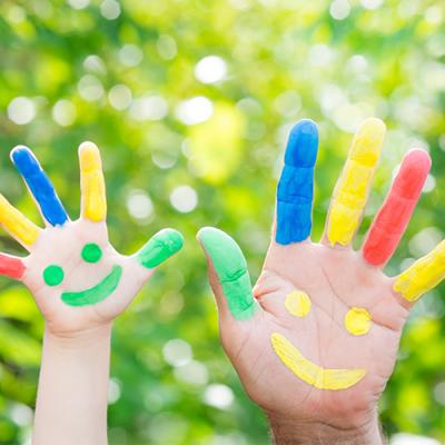 A child's hand and an adult's hand, both painted in bright colors along each finger with a smiley face on the palms